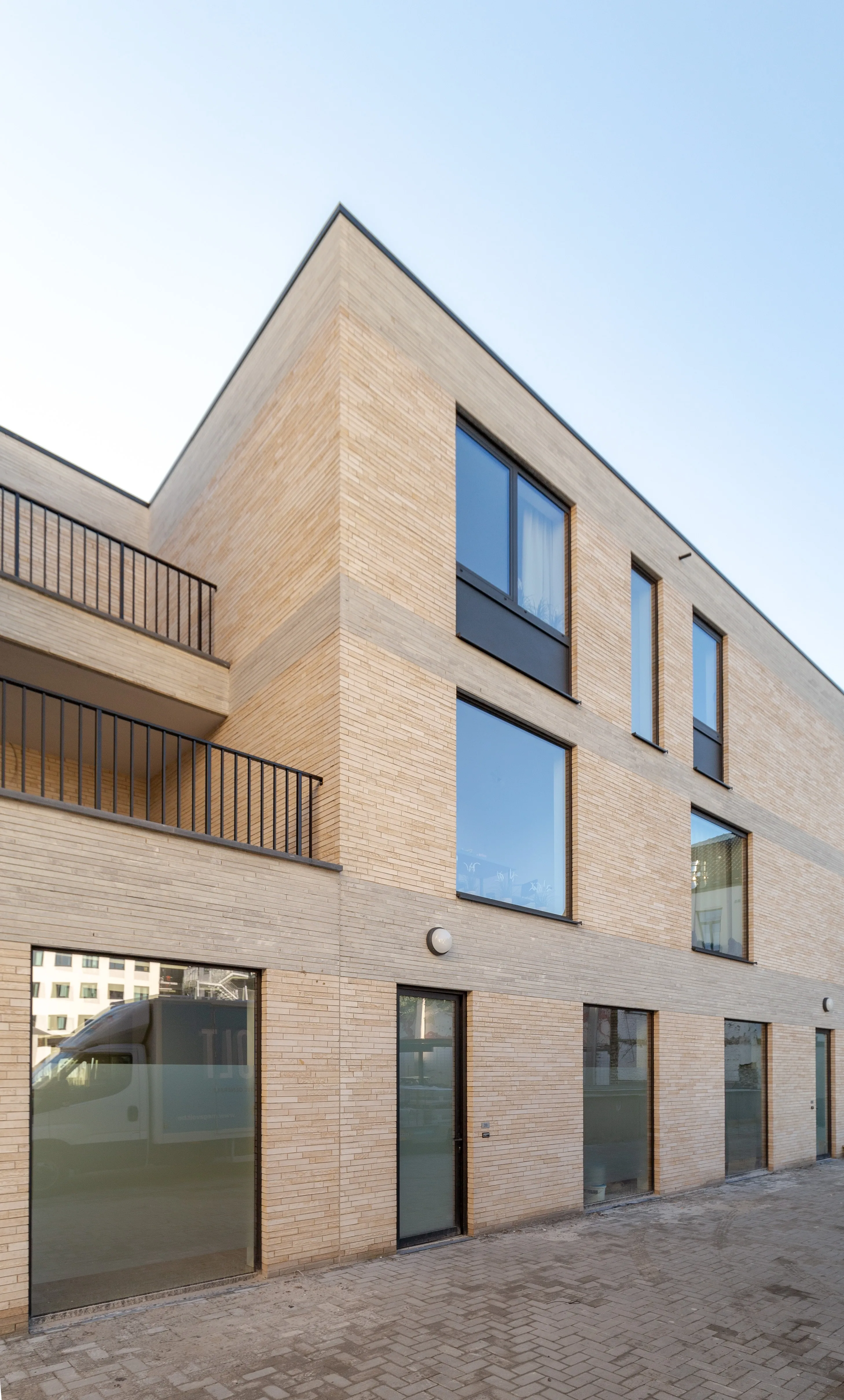 Een modern drielaags gebouw met lichte bakstenen gevel en grote rechthoekige ramen. Het gebouw heeft zwarte balkonleuningen op de eerste verdieping en glazen deuren op de begane grond. De architectuur is minimalistisch met strakke lijnen en een beige kleurenpalet. Het gebouw staat aan een geplaveide straat of binnenplaats.